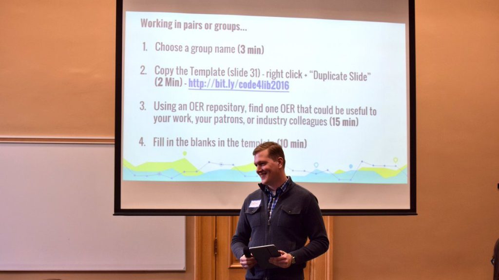 A picture of Erik in front of a projector display at a conference holding a microphone and iPad